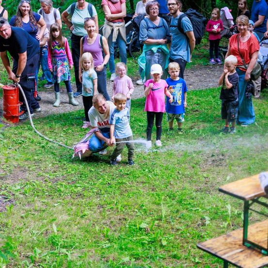 Eine Gruppe von Erwachsenen und Kindern versammelt sich auf einer Rasenfläche, wo ein Mann Wasser mit einem Schlauch versprüht. Einige Kinder stehen, während andere auf dem Boden sitzen. Im Hintergrund befinden sich Holztische.