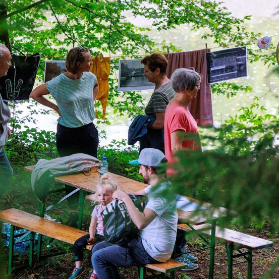 Mehrere Personen sitzen auf einer Bank in einem Park, mit einigen Kleidern, die an einem Seil hinter ihnen hängen. Ein Mädchen sitzt auf dem Schoß eines Mannes, und eine weitere Person steht in der Nähe des Tisches.