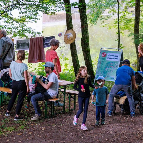 Eine Familie mit Kindern sitzt an einem Picknicktisch in einem Park, mit einer Karte und einem Schild in der Nähe. Ein erwachsener Mann schiebt einen Kinderwagen, und ein anderer Mann spricht mit einer Frau.