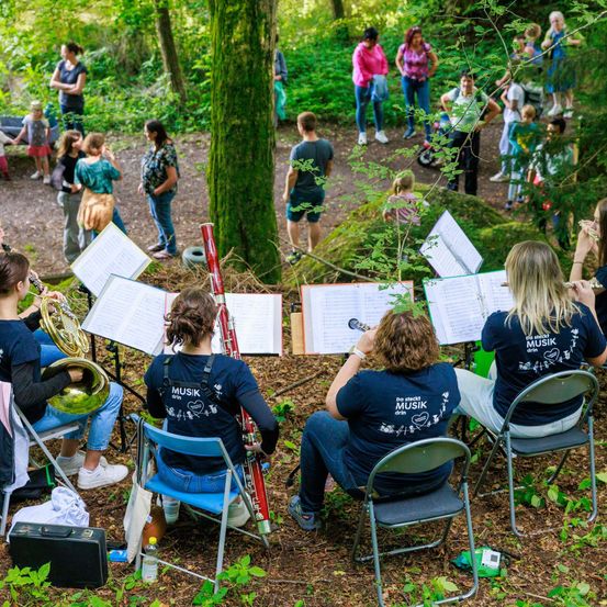 Eine Gruppe von Musikern in einem Wald, einige sitzen auf Stühlen, während andere stehen, alle tragen T-Shirts mit der Aufschrift 'Musik'. Sie sind umgeben von Notenblättern und Instrumenten wie Trompeten und Tubas.