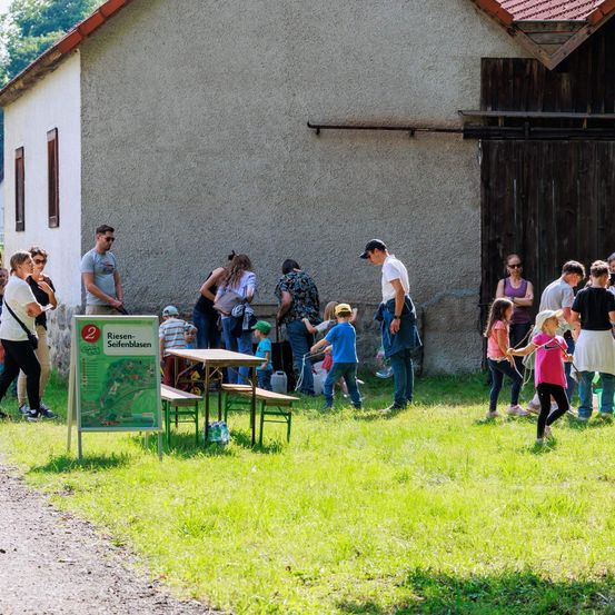 Eine Gruppe von Menschen steht vor einem Gebäude, einige gehen und andere stehen. Es gibt Kinder, die auf dem Gras spielen. Ein Schild mit einer Karte ist auf dem Boden platziert.