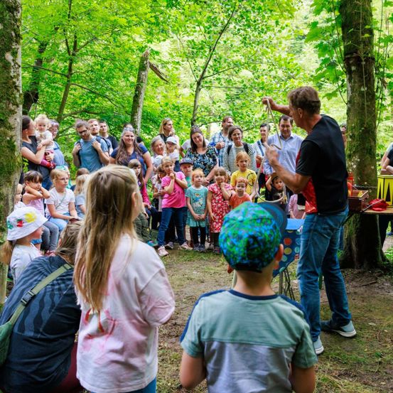 Ein Mann führt in einem Wald vor einer Gruppe von Kindern und Erwachsenen auf. Der Mann hält eine Schnur, während die Kinder zusehen. Die Kinder tragen bunte Kleidung. Einige Erwachsene halten Babys. Das Gebiet ist von Bäumen umgeben.