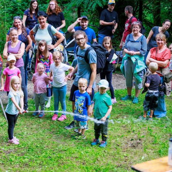 Eine Gruppe von Erwachsenen und Kindern in einem Wald, einige halten Wasserpistolen. Ein Mädchen sprüht Wasser. Sie stehen auf saftigem Boden mit Bäumen im Hintergrund.