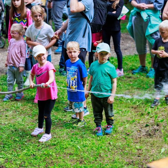 Eine Gruppe von Kindern hält einen langen Schlauch, der Wasser sprüht, während andere sie beobachten. Die Kinder stehen auf einem Grasfeld.