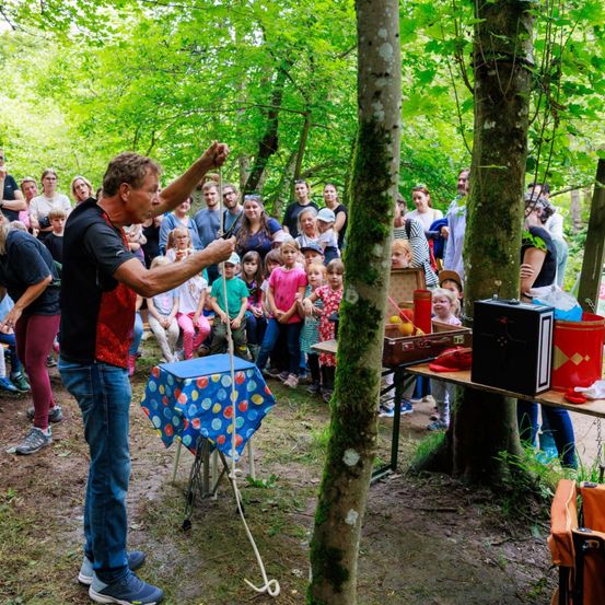 Ein Mann führt einen Zaubertrick im Wald für eine Gruppe von Kindern und Erwachsenen vor. Es gibt Tische mit Gegenständen und einen roten Trommel auf dem Boden.