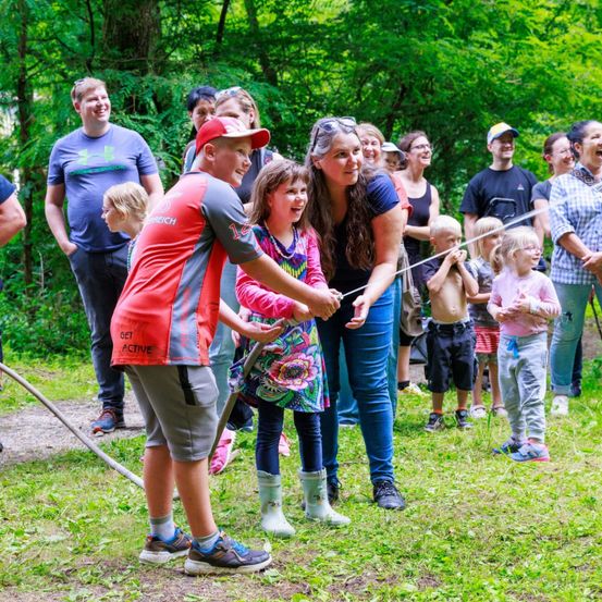 Eine Gruppe von Menschen in einem Wald, ein Mann sprüht Wasser auf eine Frau und ein Mädchen, während andere zusehen und lachen.