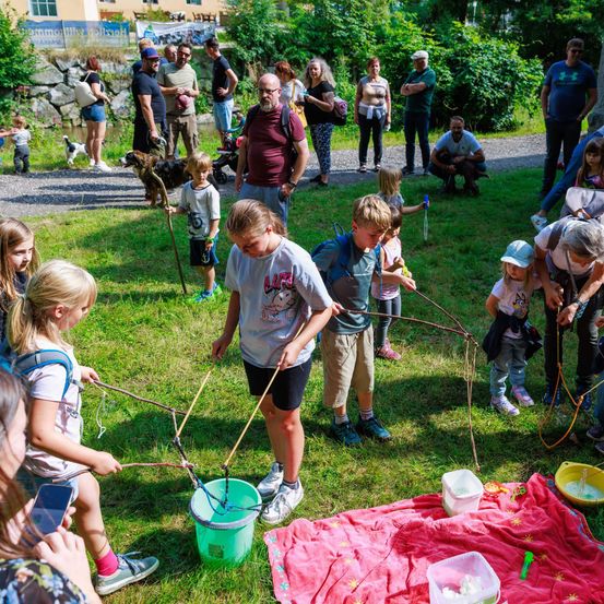 Eine Gruppe von Kindern und Erwachsenen versammelt sich auf einem Rasenfeld. Einige benutzen Angelruten und es gibt Eimer mit Wasser. Menschen stehen herum und beobachten.