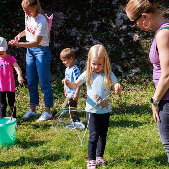 Eine Familie mit drei Kindern, zwei Erwachsenen und einem Eimer steht auf einer Wiese. Ein Kind hält eine Seifenblasenwand.