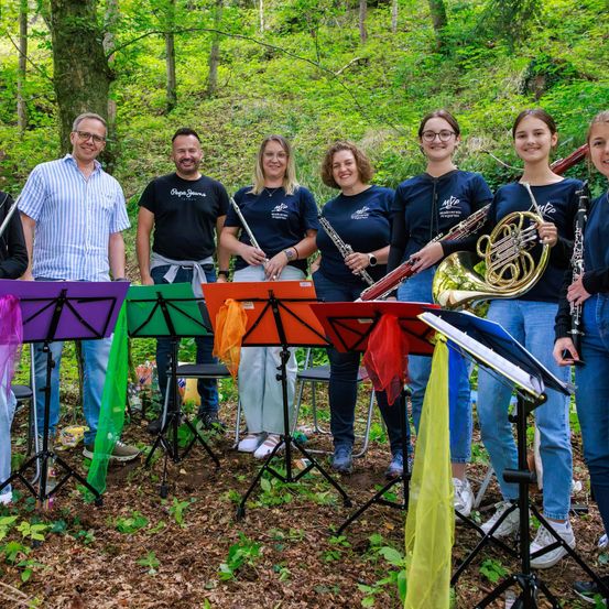 Eine Gruppe von Musikern, darunter drei Frauen und zwei Männer, steht in einem Wald mit Musikinstrumenten in den Händen, wahrscheinlich in Vorbereitung auf eine Aufführung.
