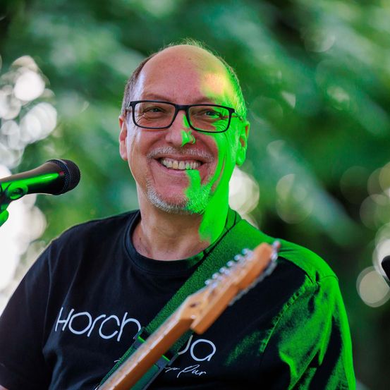 Ein Mann mit einer schwarzen T-Shirt und grüner Farbe im Gesicht spielt Gitarre. Er lächelt und steht vor einem Mikrofon.