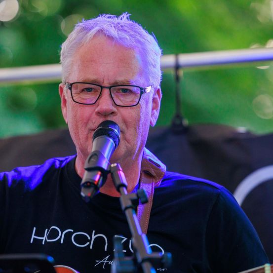 Ein Mann mit weißen Haaren und Brille singt in ein Mikrofon. Er trägt ein schwarzes T-Shirt mit weißem Text. Dahinter befindet sich ein schwarzer Hintergrund mit einem weißen Symbol.