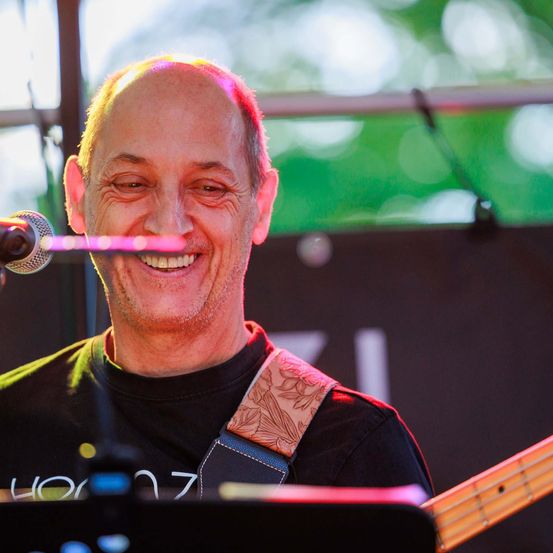 Ein Mann mit einem Gitarrengurt lächelt in ein Mikrofon, möglicherweise bei einer Bühnenperformance.
