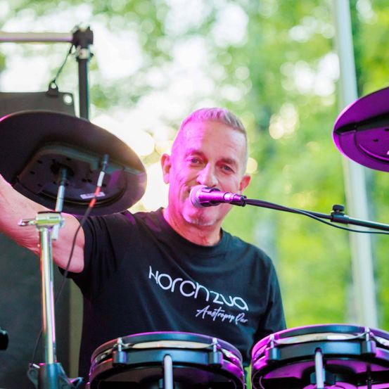 Ein Mann mit kahlem Kopf trägt ein schwarzes T-Shirt mit weißen Schriftzügen. Er steht vor einem Mikrofon und hält einen Drumstick in der rechten Hand. Vor ihm stehen Trommeln.