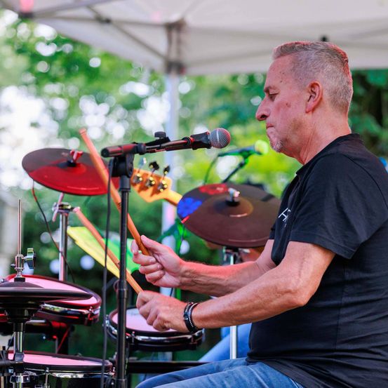 Ein älterer Mann spielt die Drums auf einer Bühne mit Bäumen im Hintergrund.