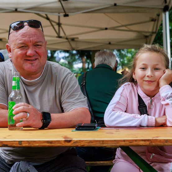 Ein Mann und ein junges Mädchen sitzen an einem Tisch unter einem Zelt und lächeln in die Kamera. Der Mann hält eine Flasche. Ein anderer sitzt hinter ihnen.