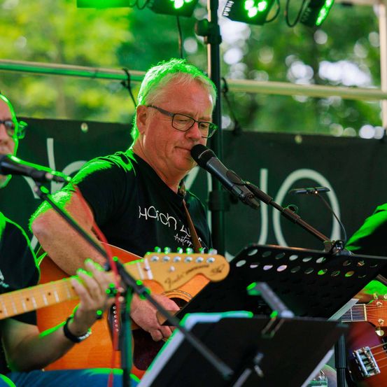 Ein Mann mit Brille und einem grünen T-Shirt spielt Gitarre und singt in ein Mikrofon. Eine andere Person spielt Gitarre an der Seite. Die Bühne hat grüne Lichter und ein Banner dahinter.