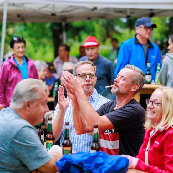 Im Freien versammelte Menschen klatschen, ein Mann raucht und Bierflaschen stehen auf dem Tisch unter einem weißen Zelt.