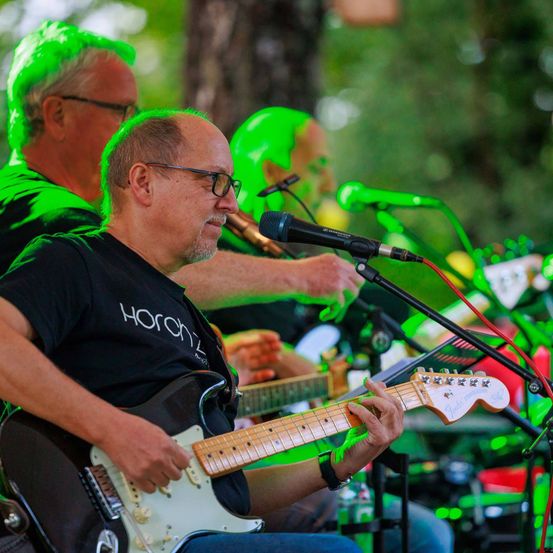 Ein Mann spielt Gitarre auf einer Bühne, mit einem Scheinwerfer auf ihm. Ein anderer Mann spielt Flöte hinter ihm. Sie sind umgeben von einem Mikrofon und anderen Instrumenten.