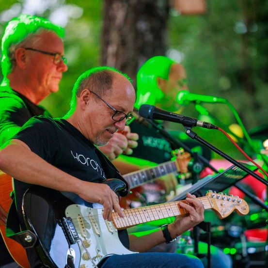Drei Männer spielen Gitarren auf der Bühne. Einer greift die Gitarre, während die anderen beiden singen. Mikrofone stehen vor ihnen.