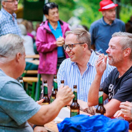Eine Gruppe von Erwachsenen, darunter Männer mit Brille, sitzt um einen Tisch mit Bierflaschen herum. Sie scheinen eine zwanglose Zusammenkunft in einem Außenbereich zu genießen.