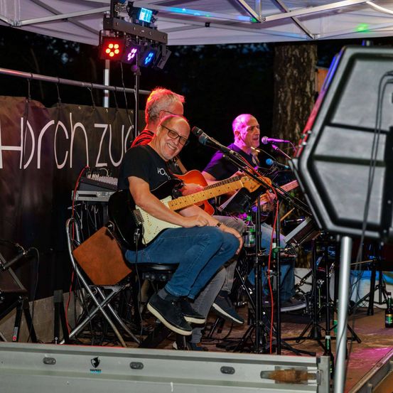 Drei Musiker treten bei Nacht auf der Bühne auf. Der Mann in der Mitte spielt Gitarre und lächelt. Die anderen spielen ebenfalls Gitarren und singen in Mikrofone. Ausrüstung und Flaschen sind auf der Bühne.