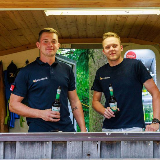 Zwei Männer stehen hinter einer Bartheke und halten jeweils eine Flasche Freistadl-Bier. Sie tragen passende blaue Uniformen mit Logos, einer mit Poloshirt und der andere mit T-Shirt. Der Schauplatz ist im Freien, mit einer Holzdecke und einem Coca-Cola-Kühlschrank hinter ihnen.