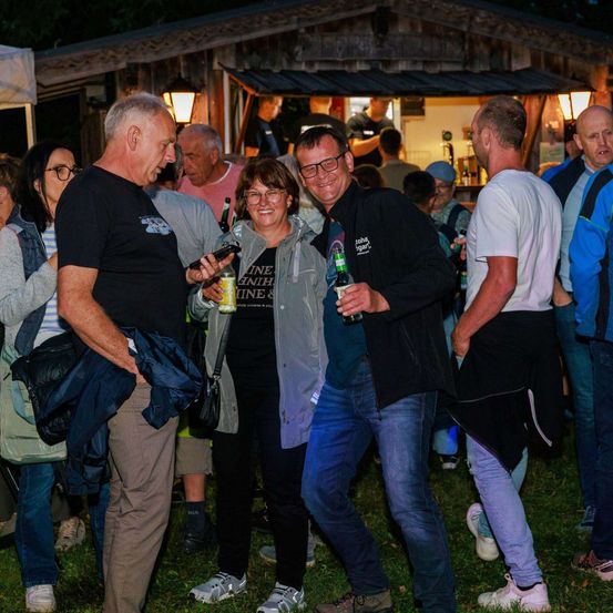 Eine Gruppe von Erwachsenen steht bei Nacht draußen, lächelt und hält Getränke in den Händen. Im Hintergrund befindet sich eine Holzbar und ein Essbereich im Freien.