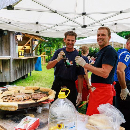 Zwei Männer in Schürzen stehen unter einem Zelt an einem Tisch, lächeln und halten Flaschen Bier in den Händen. Sie bereiten Essen zu, darunter Würste auf einem großen Grill. Auf dem Tisch befindet sich eine Plastiktüte mit Brötchen, eine Schachtel und eine Flasche. Im Hintergrund sind weitere Menschen, eine Holzstruktur und Bäume zu sehen.