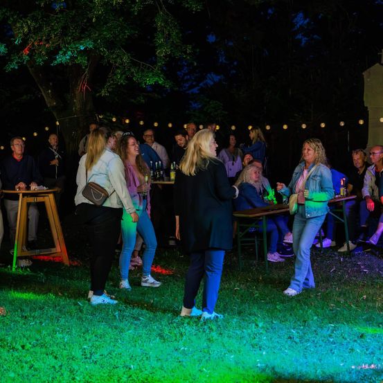 Eine Gruppe von Menschen trifft sich bei einer nächtlichen Outdoor-Veranstaltung, die von grünen und roten Lichtern beleuchtet wird. Sie versammeln sich um Tische und stehen auf einer Rasenfläche.