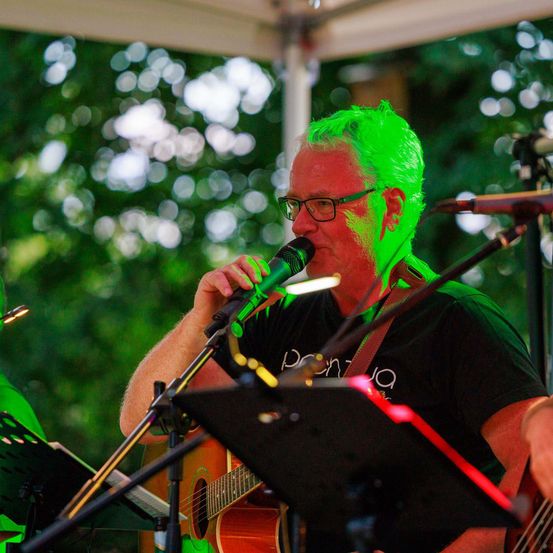 Ein Mann mit grünen Haaren singt in ein Mikrofon, während er eine Gitarre unter einem Baldachin im Freien spielt.