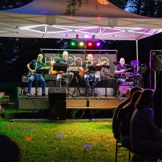 Eine Band aus vier Männern spielt auf einer Bühne unter einem Zelt bei Nacht, mit bunten Lichtern und einem Banner. Zuschauer sitzen auf dem Gras und schauen zu.