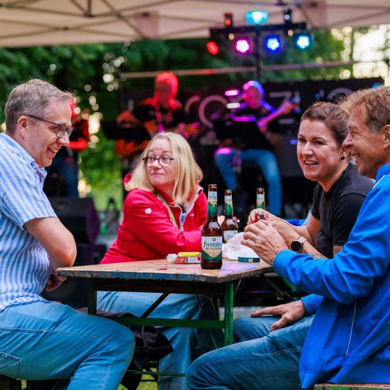 Vier Personen sitzen mit Bierflaschen an einem Tisch und lächeln. Eine Band spielt im Hintergrund.