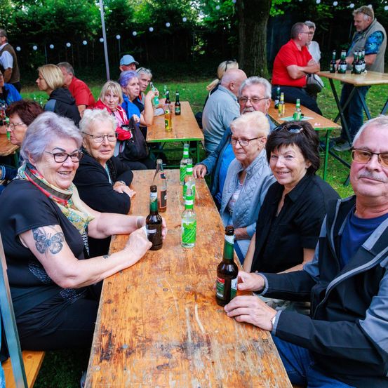 Eine Gruppe von Menschen sitzt an einem langen Holztisch in einem Außenbereich, lächelt und hält Flaschen. Hinter ihnen sitzen weitere Personen an anderen Tischen. Der Bereich ist von Bäumen umgeben und hat hängende Lichter.
