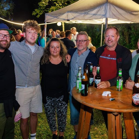 Eine Gruppe von Erwachsenen lächelt und posiert für ein Foto bei einer nächtlichen Outdoor-Veranstaltung mit Flaschen auf einem Tisch und einem Zelt im Hintergrund.