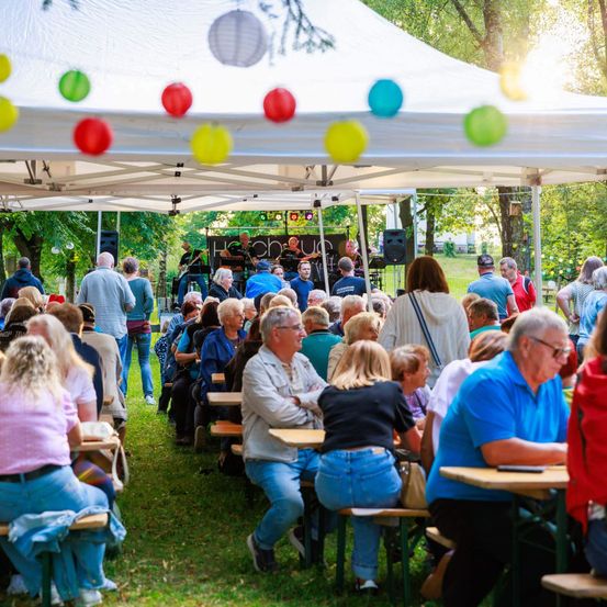 Eine Gruppe von Menschen versammelt sich unter einem Zelt für eine musikalische Veranstaltung. Eine Band spielt auf der Bühne, während die Besucher an Tischen sitzen, einige zuschauen und andere sich unterhalten. Bunte Papierlaternen hängen am Zelt.
