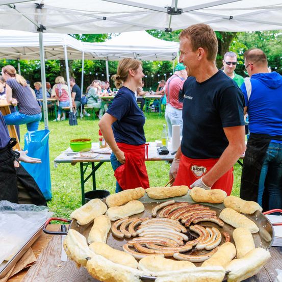 Ein Mann und eine Frau in Schürzen servieren Essen im Freien. Der Mann lacht, während die Frau ihn ansieht. Viele Menschen sitzen um Tische unter weißen Zelten.