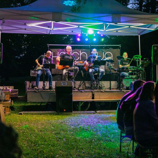 Eine Gruppe Musiker tritt auf einer Bühne unter einem Zelt mit farbigen Lichtern, Gitarren und Trommeln auf, während ein Publikum zusieht.