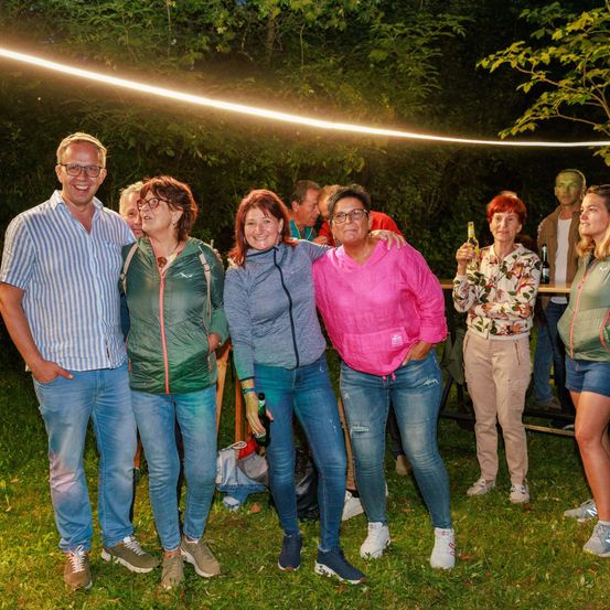 Eine Gruppe von Menschen steht auf einem Grasfeld und lächelt, während sie Getränke in der Hand halten. Im Hintergrund sind Bäume und ein beleuchteter Weg zu sehen.