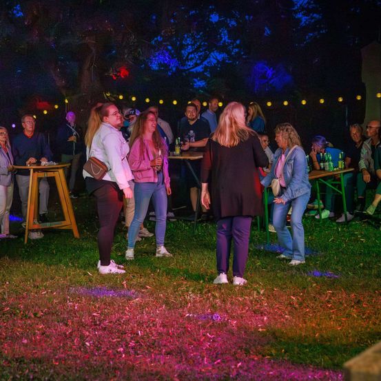 Eine Gruppe von Menschen versammelt sich bei Nacht auf einem Rasenfeld, das von farbigen Lichtern beleuchtet wird. Einige stehen, andere sitzen auf Bänken. Es gibt Tische mit Flaschen und Gläsern. Bäume stehen dahinter, ebenfalls beleuchtet.