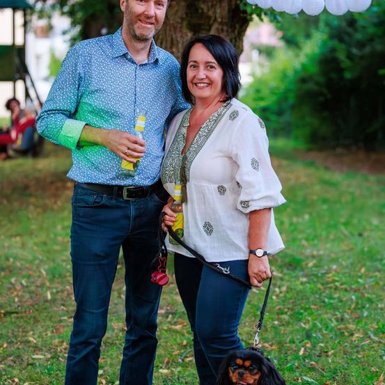 Ein Mann und eine Frau stehen lächelnd für die Kamera auf einem Rasen. Jeder hält eine Flasche und die Frau hält auch einen Hund an der Leine.