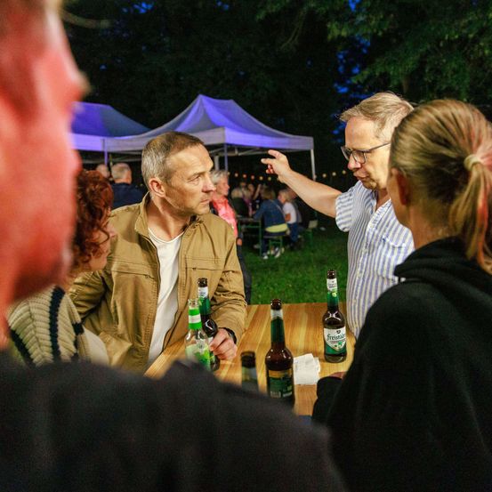 Mehrere Erwachsene versammeln sich um einen Tisch mit Bierflaschen und einem Mann, der zeigt. Im Hintergrund befinden sich Zelte und weitere Menschen.