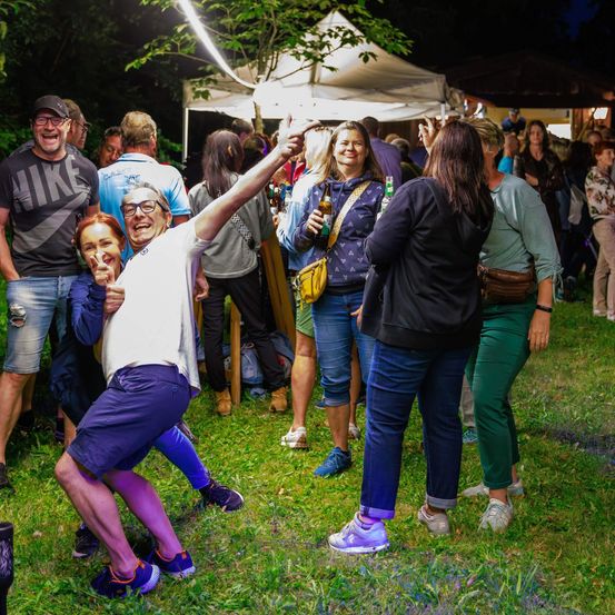 Eine Gruppe von Menschen tanzt im Freien. Ein Mann in einem weißen T-Shirt hebt die Hände. Eine Frau in der Mitte hält eine Flasche. Ein Zelt steht hinter ihnen.