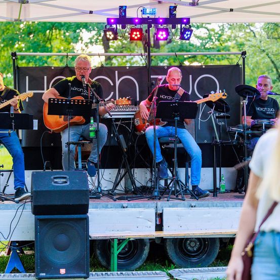 Eine Band tritt auf einer Bühne unter einem Zelt auf. Vier Musiker spielen Gitarren und einen Bass. Scheinwerfer sind über der Bühne montiert. Ein Publikum beobachtet von der Seite.