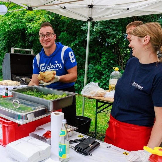 Ein Mann in einem Carlsberg-Shirt hält ein Sandwich und steht unter einem Zelt mit Essensbehältern. Eine Frau mit einem roten Schürze spricht mit ihm. Hinter ihnen steht ein Tisch mit einer Flasche und einer Aufbewahrungsbox. Bäume und ein Grill sind im Hintergrund.