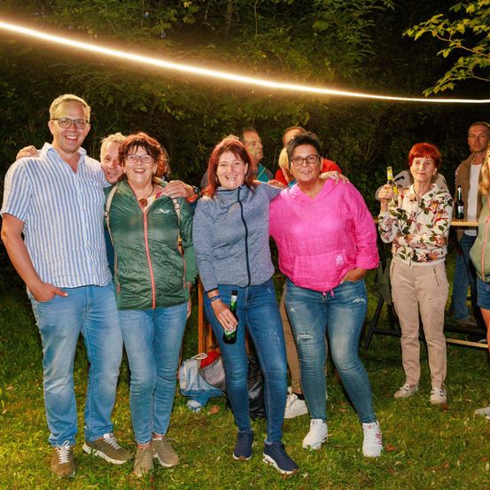 Eine Gruppe von Menschen steht auf einer Rasenfläche und posiert für ein Foto. Sie tragen Freizeitkleidung, und einige halten Flaschen. Der Bereich ist von Bäumen umgeben, und es gibt einen Tisch mit Flaschen und anderen Gegenständen.