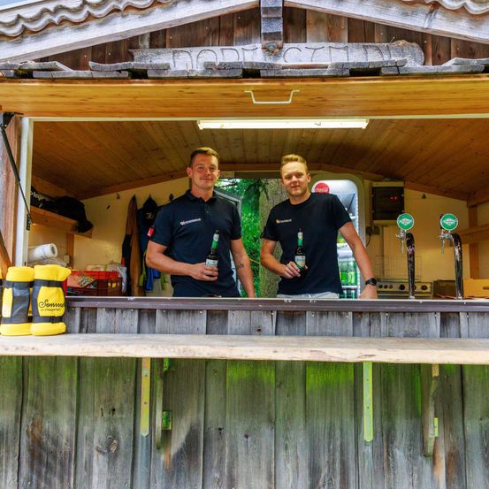 Zwei Männer stehen hinter einer Bartheke in einer Holzhütte und halten Flaschen. Sie tragen passende Uniformen und lächeln. Dahinter steht ein Coca-Cola-Kühler und ein Baum.