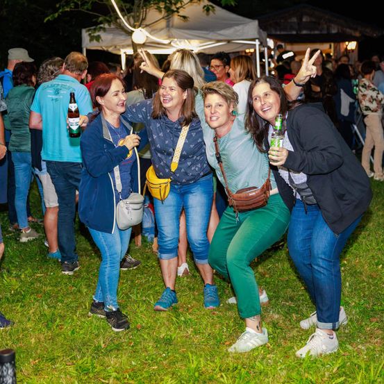 Fünf Frauen in Freizeitkleidung posieren für ein Foto bei einer nächtlichen Outdoor-Veranstaltung, lächeln und halten Getränke in den Händen.
