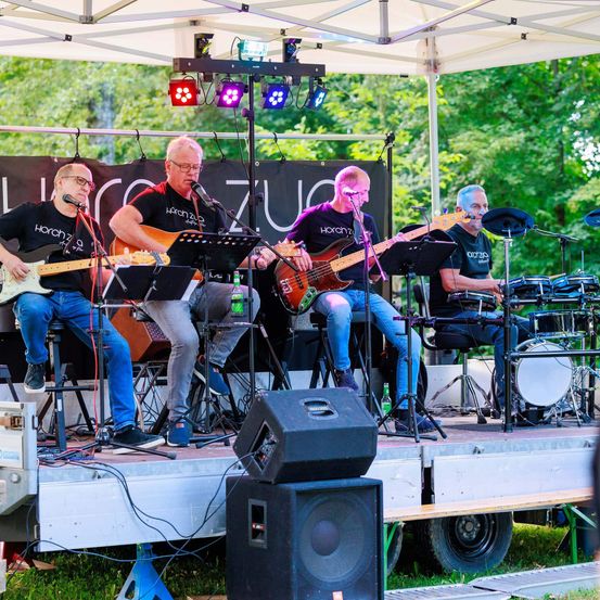 Fünf Musiker spielen auf einer Freiluftbühne unter einem weißen Zelt. Zwei spielen Gitarre, zwei spielen Bass und einer spielt Schlagzeug. Im Hintergrund befinden sich Bäume und ein Schild mit der Aufschrift 'Horch zu'.