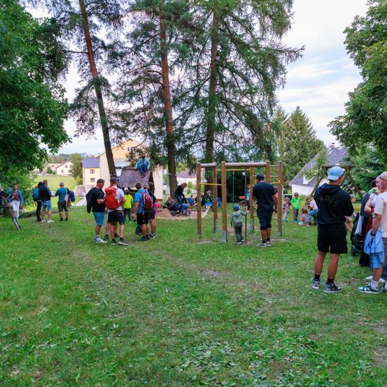 Eine Gruppe von Menschen versammelt sich in einem Park mit Bäumen und Spielplatzgeräten. Einige stehen, andere sitzen oder gehen. Eine Person sitzt auf einem Ast.