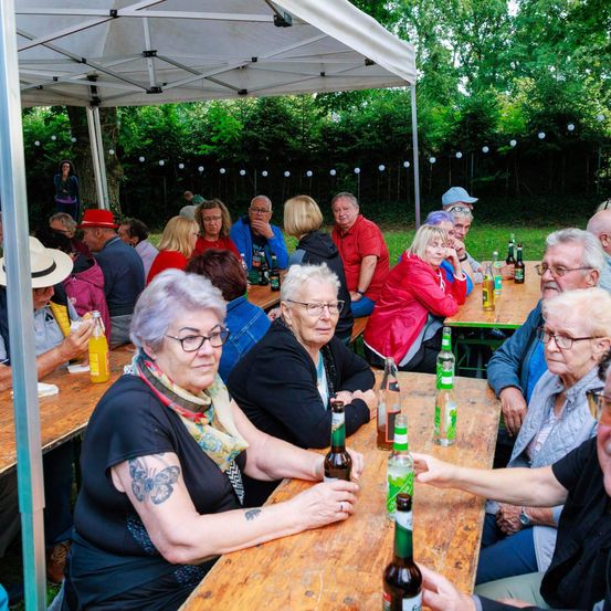 Eine Gruppe älterer Menschen versammelt sich an einem Picknicktisch im Freien unter einem Zelt, mit Bäumen und Büschen im Hintergrund. Sie trinken Bier und unterhalten sich miteinander.